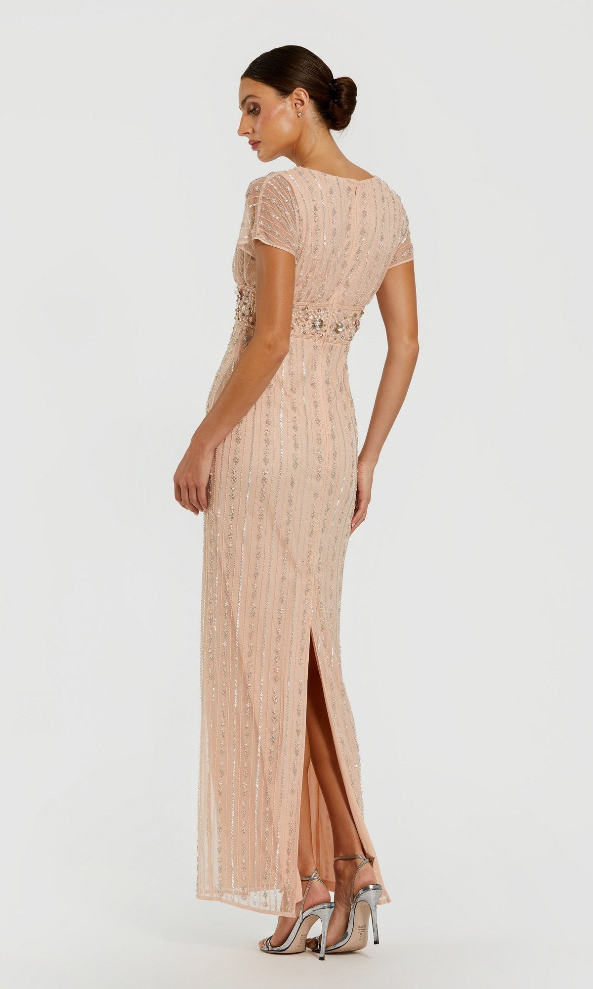 A woman stands against a plain background wearing the Mac Duggal 94147 long formal dress—a light pink, short-sleeve gown featuring beaded pearl embellishments and a back slit—paired with high-heeled sandals.